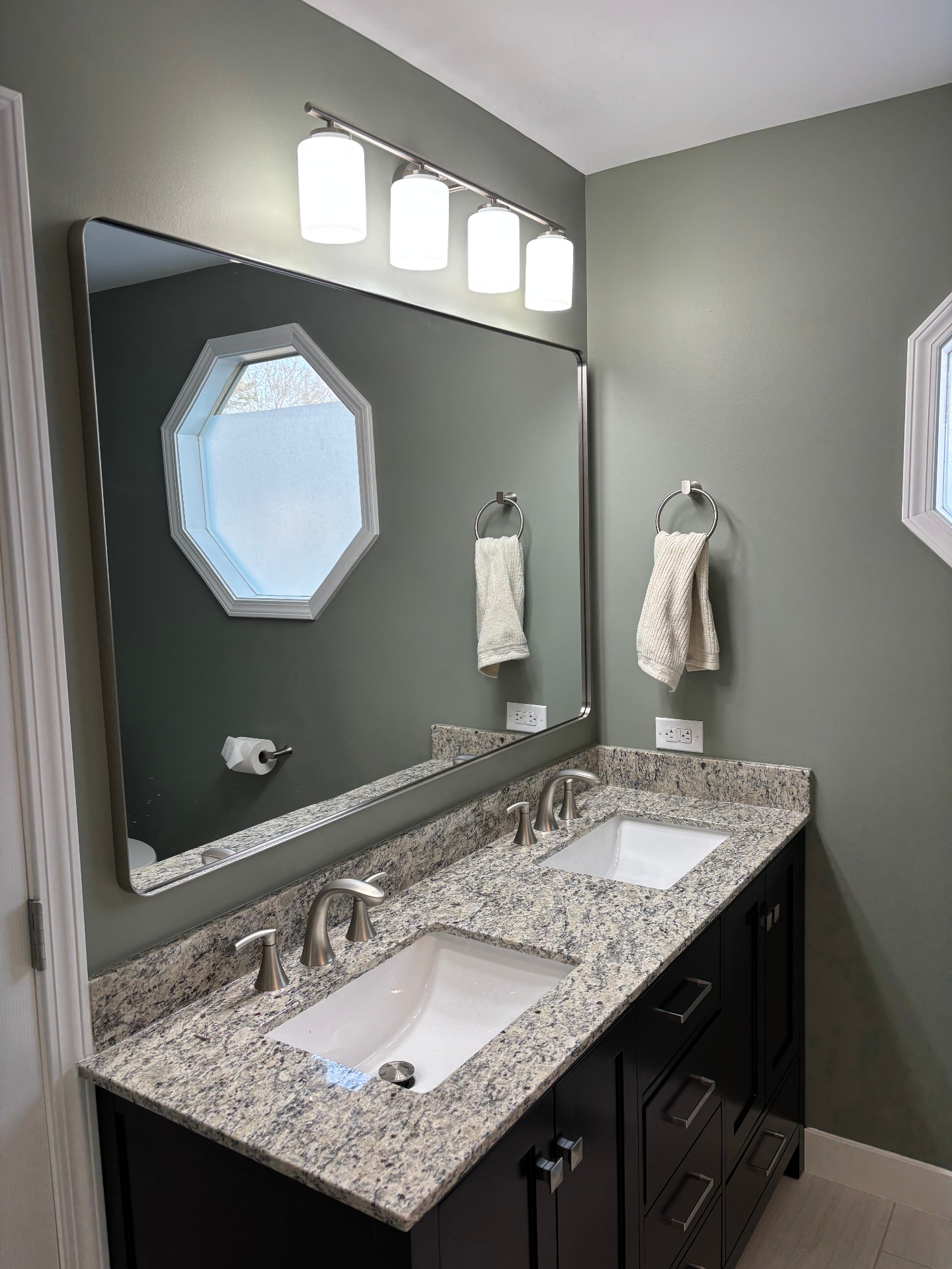Vanity area with sage walls, granite double-sink top, framed mirror and four-light brushed-nickel vanity fixture